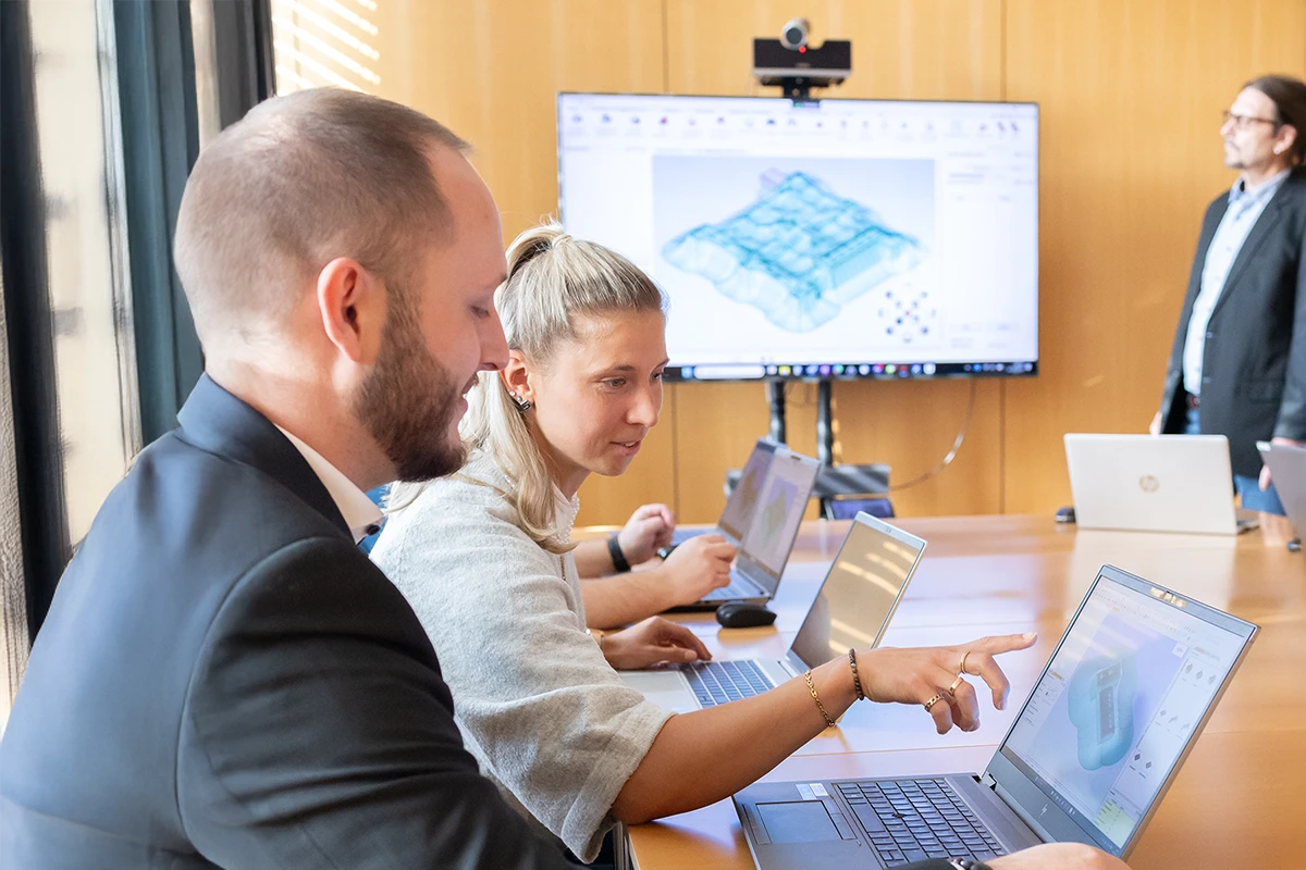 Three people working on laptops in a meeting room while a presenter stands near a large screen displaying a 3D model.