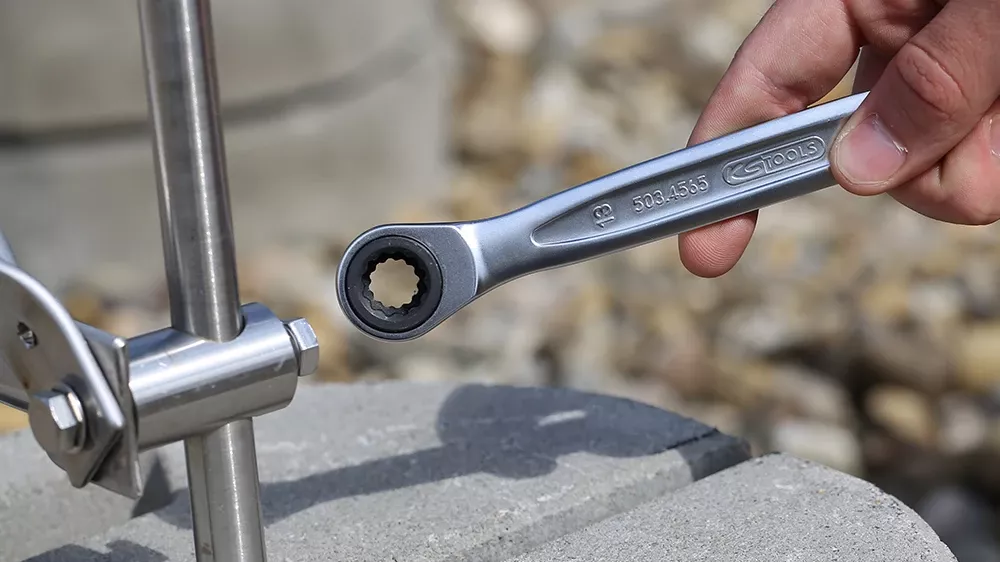 Hand using a ratcheting wrench to tighten a clamp on a vertical metal rod fixed into a concrete block.