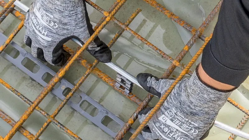 Gloved hands attach a metal connector to round steel rebars on a wet rebar grid, viewed from above.