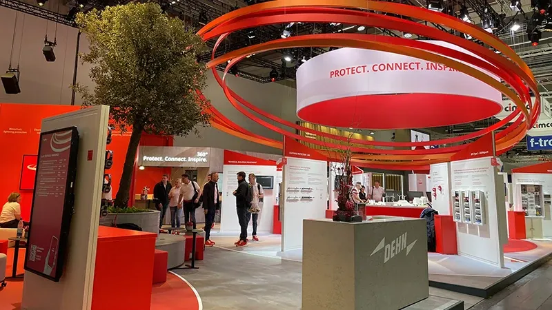 Exhibition booth with red and white design, several people, large red rings above the booth, and a tree in a red planter