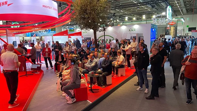 People sitting and standing in a trade show hall facing a stage with red carpet and presentation.