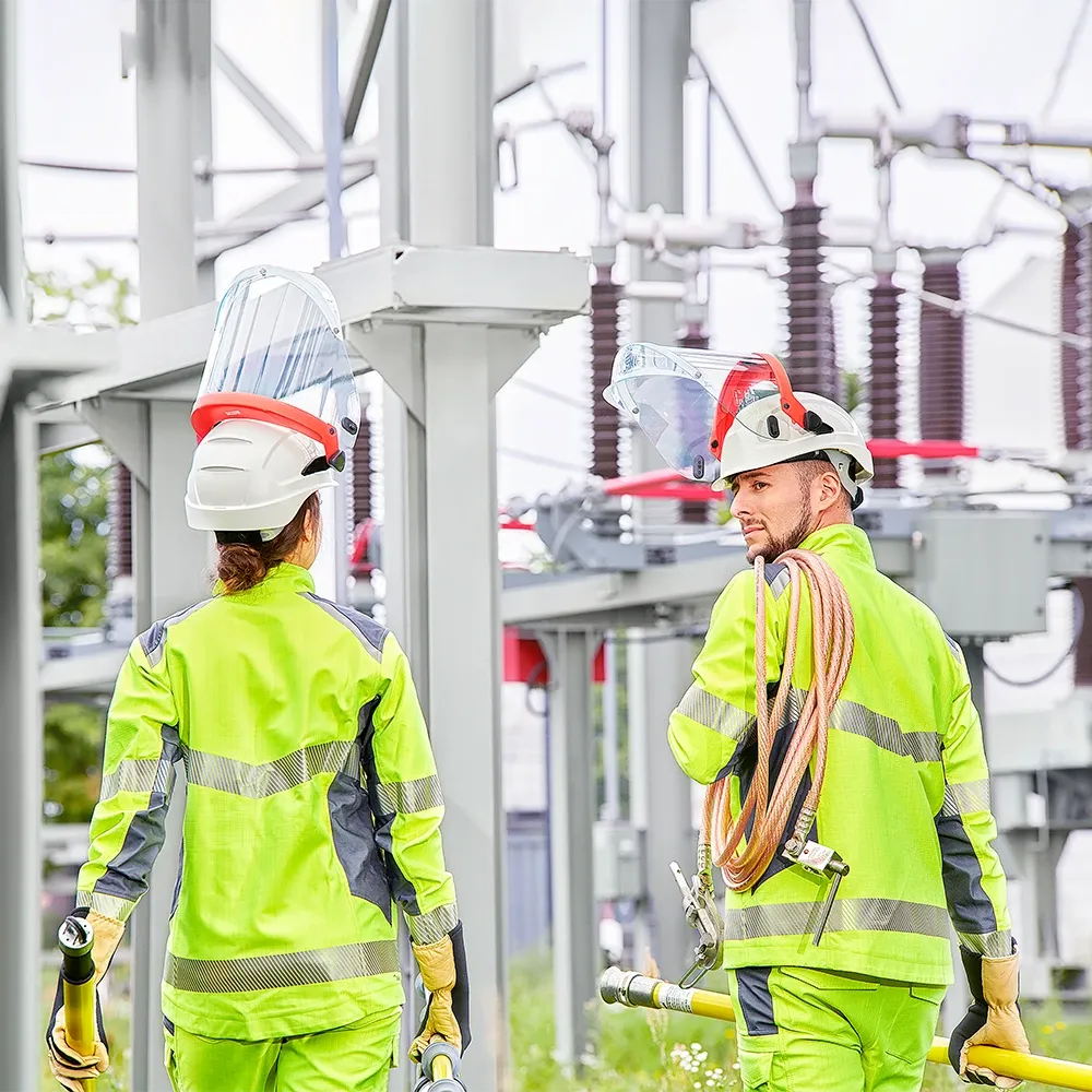 Une femme et un homme en tenue de protection complète se tiennent devant des installations dans un poste de transformation.