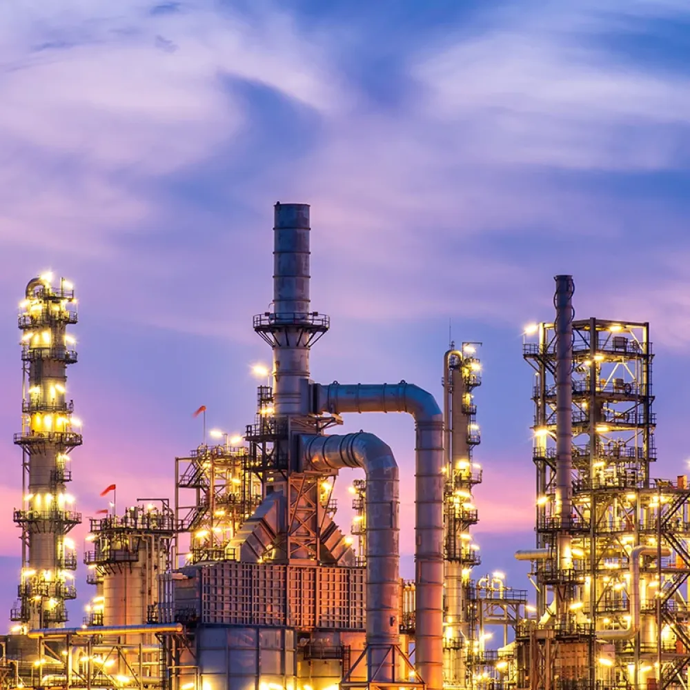 Beleuchtete Chemieanlage im Abendlicht mit zahlreichen Türmen, Pipelines und Lichtern vor einem lila gefärbten Himmel.