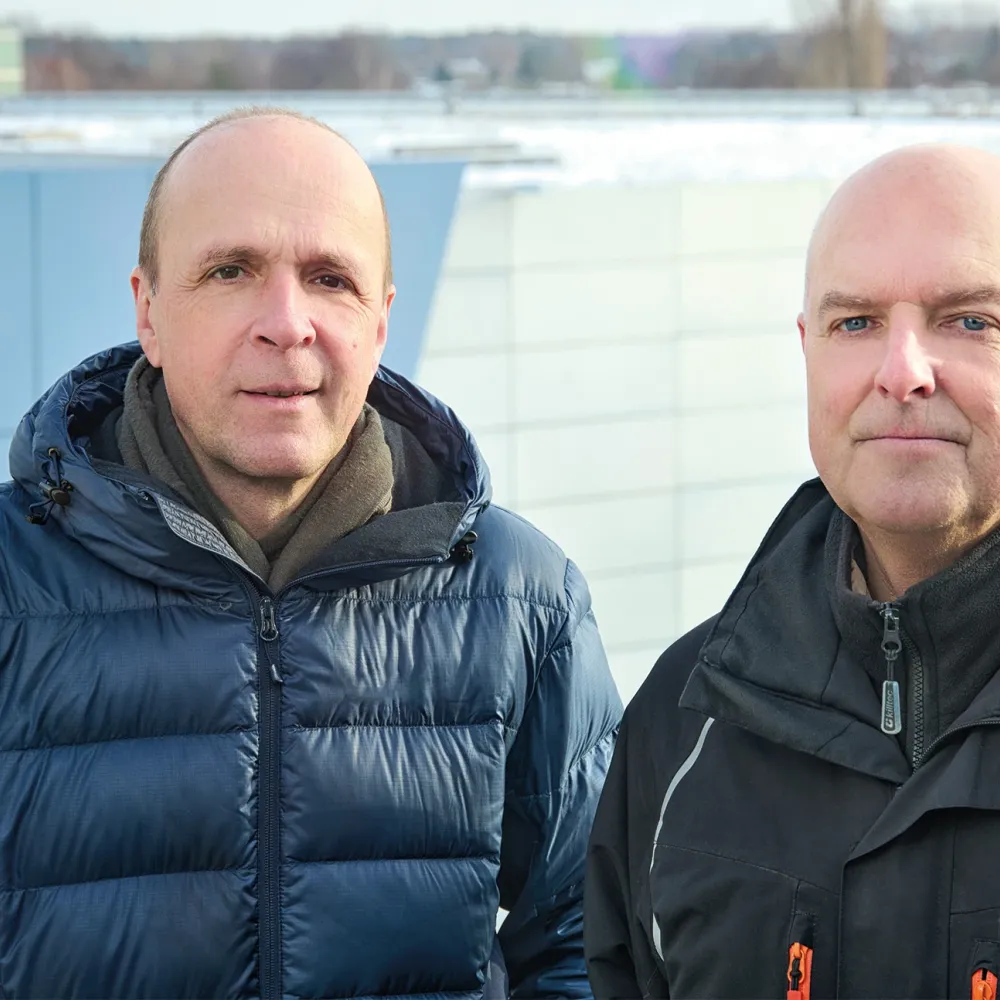 Deux hommes en veste bleue et noire devant un bâtiment industriel gris-bleu à toit plat.