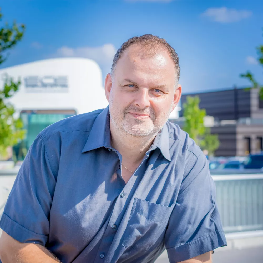 Man in a short-sleeved shirt smiling outdoors with modern buildings in the background.