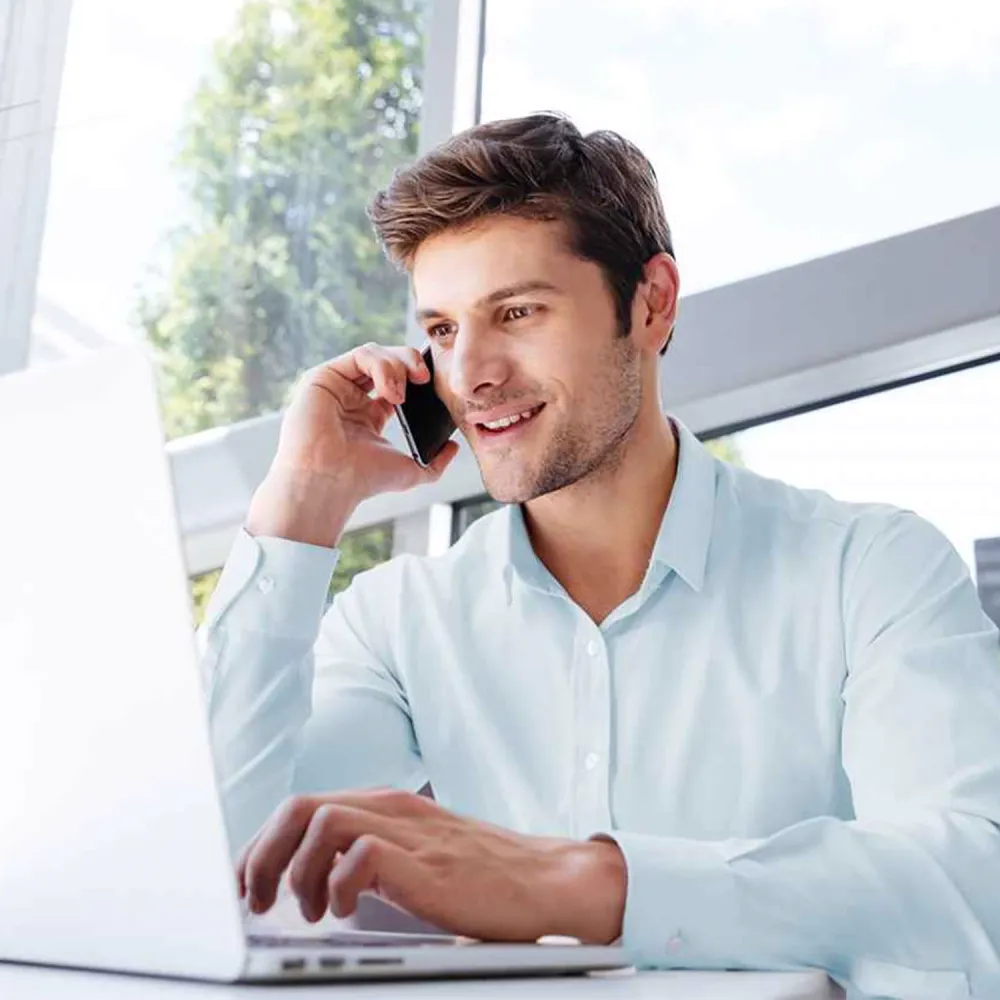 Man in a light blue shirt talking on a smartphone while working on a laptop in a bright office setting.