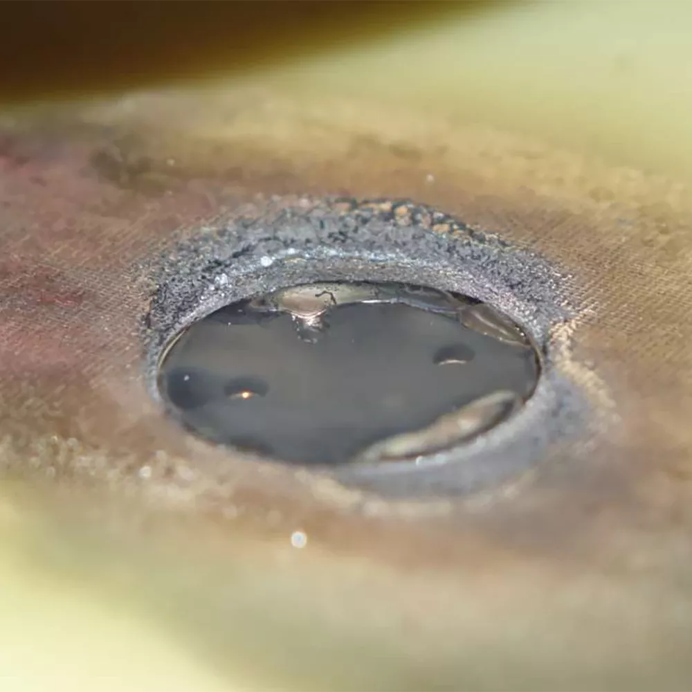 Close-up of a circular hole in a metal surface filled with liquid, showing heat discoloration and rough edges.