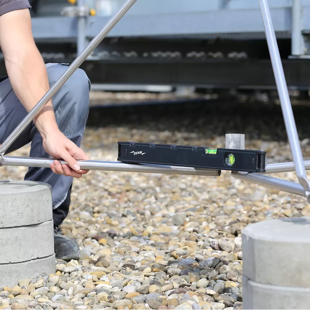 Person aligning metal rods with a spirit level on a gravel surface, hands visible near concrete blocks.