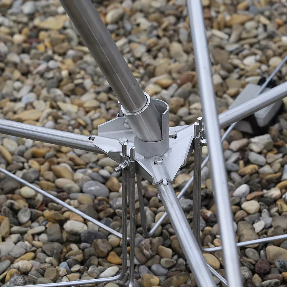 Person aligning metal rods with a spirit level on a gravel surface, hands visible near concrete blocks. Close-up of a metal joint connecting angled rods with bolts and clamps, installed on a gravel surface.