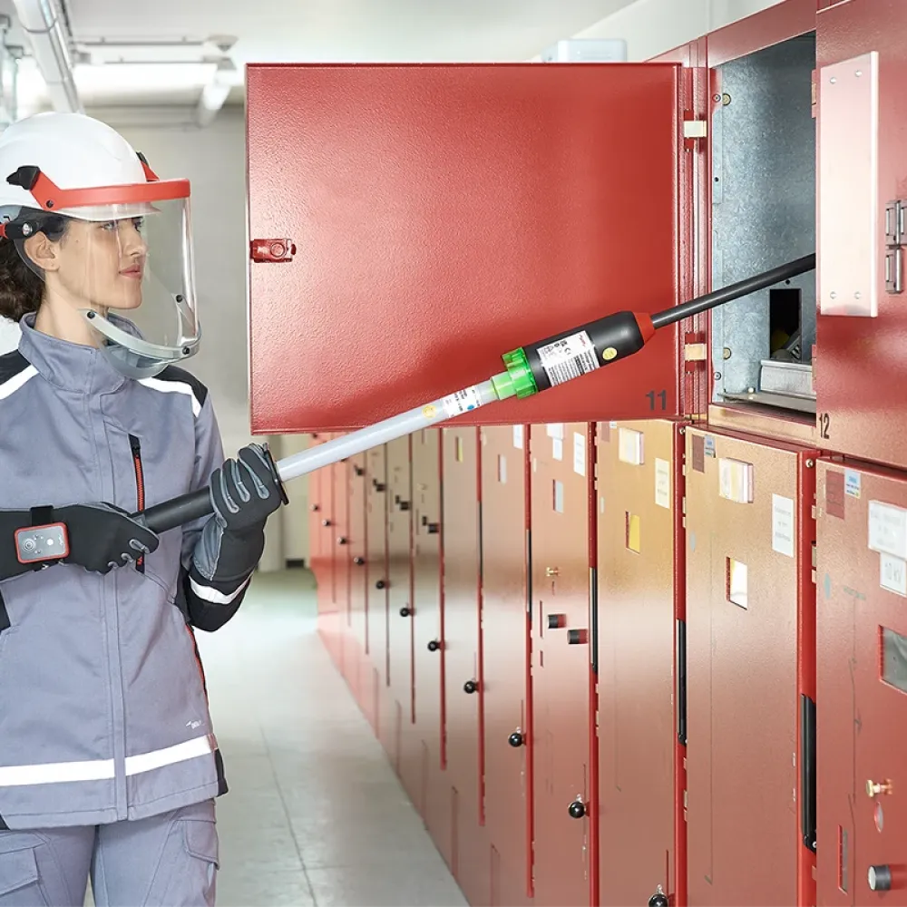Technician wearing face shield and protective clothing uses an insulated testing pole on an open red electrical switchgear cabinet.