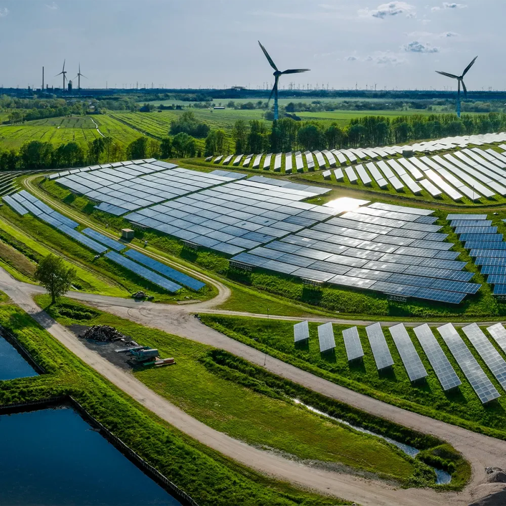 Large solar power plant with rows of photovoltaic panels across green fields, wind turbines visible in the background.