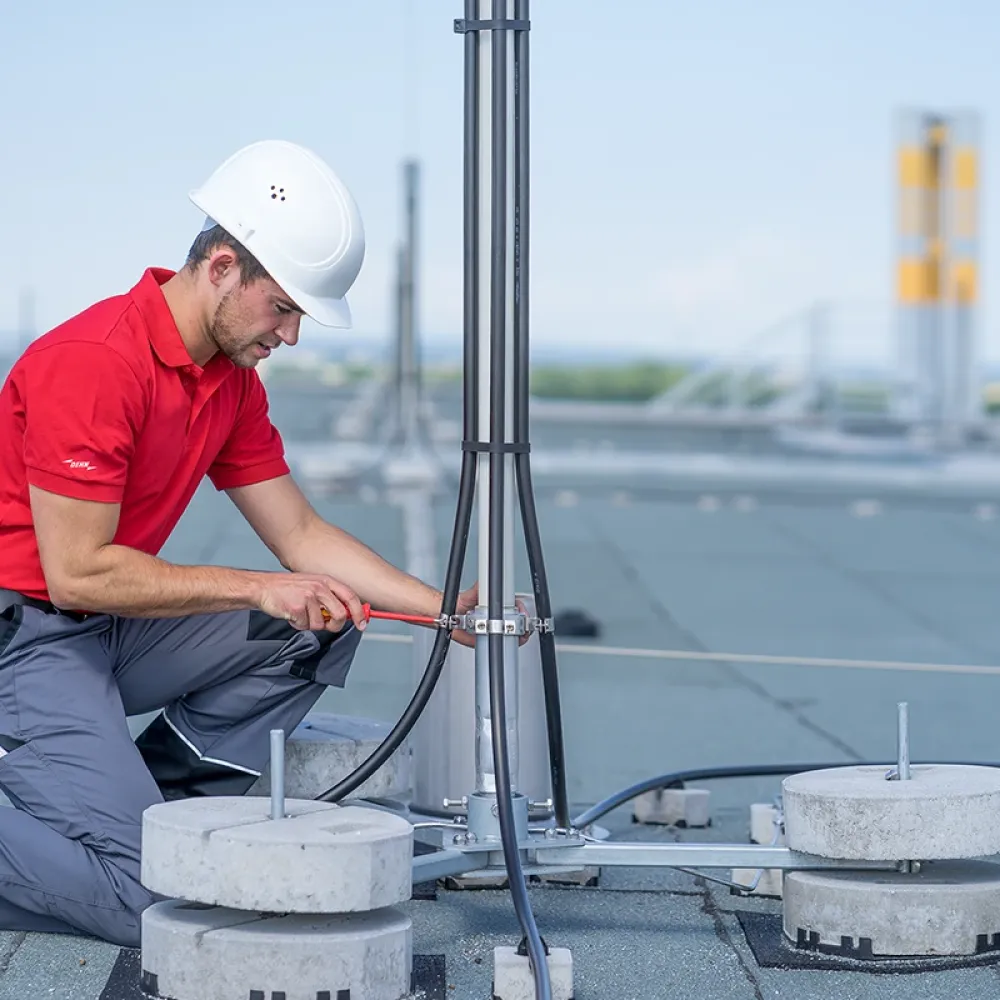 Technician in red shirt and white hard hat installing grounding cables on a rooftop lightning protection mast.