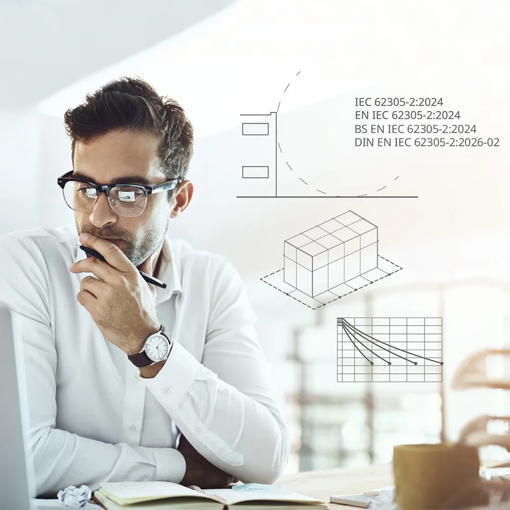 Man in glasses working at a laptop, holding a pen thoughtfully, with technical diagrams and IEC 62305-2:2024 graphics overlaid in the background.