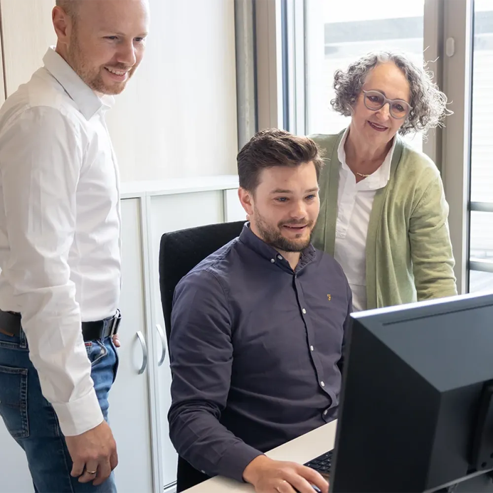 Three people at a desk in an office, two standing and one seated, looking at a computer monitor together near a window.