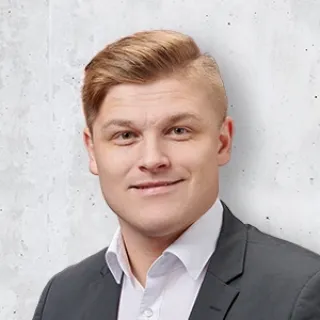 Portrait of a young man in a dark suit jacket and white shirt, smiling slightly against a light gray background.