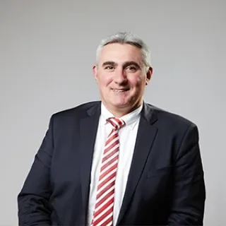 Portrait of a man in a dark suit, white shirt, and red striped tie, smiling against a plain light background.