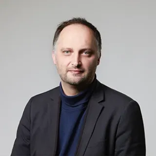 Portrait of a man with short hair wearing a dark blazer and navy top, facing the camera against a plain light background.