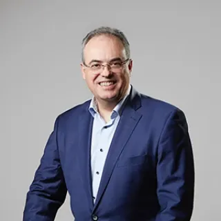 A studio portrait man wearing glasses, dressed in a dark blue suit jacket over a light shirt.