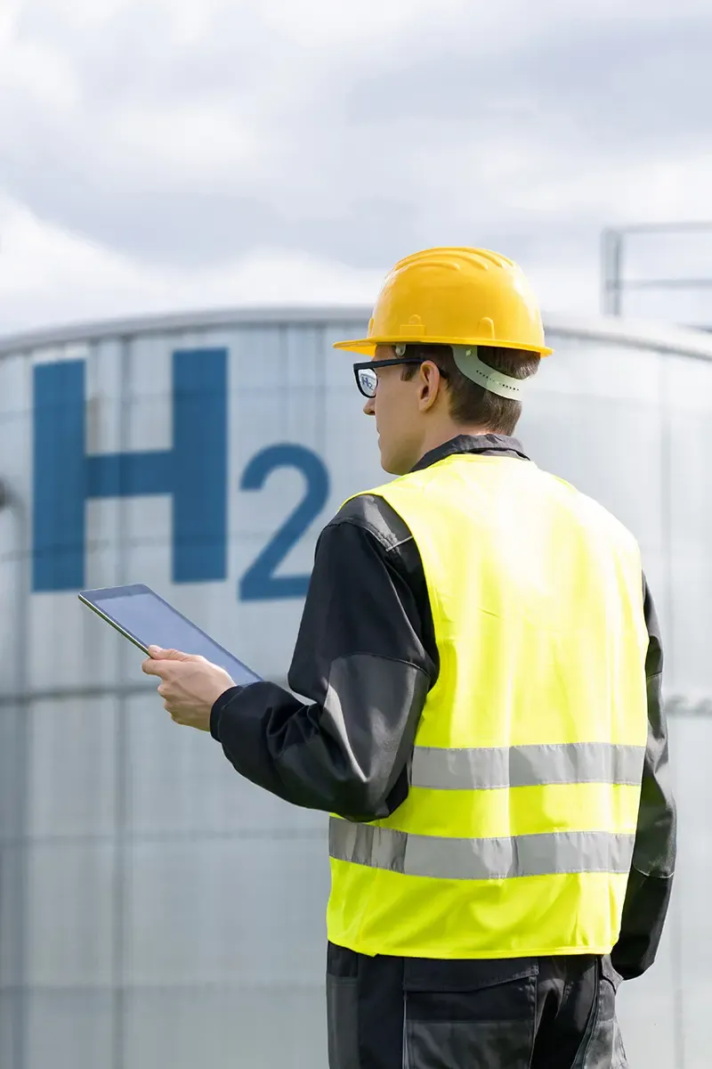 A captivating image displays a man in a yellow hard hat and vest, standing by an H2 cylinder. The images unique blend of a worker holding a tablet amidst a serene blue sky with clouds creates a peaceful construction scene.