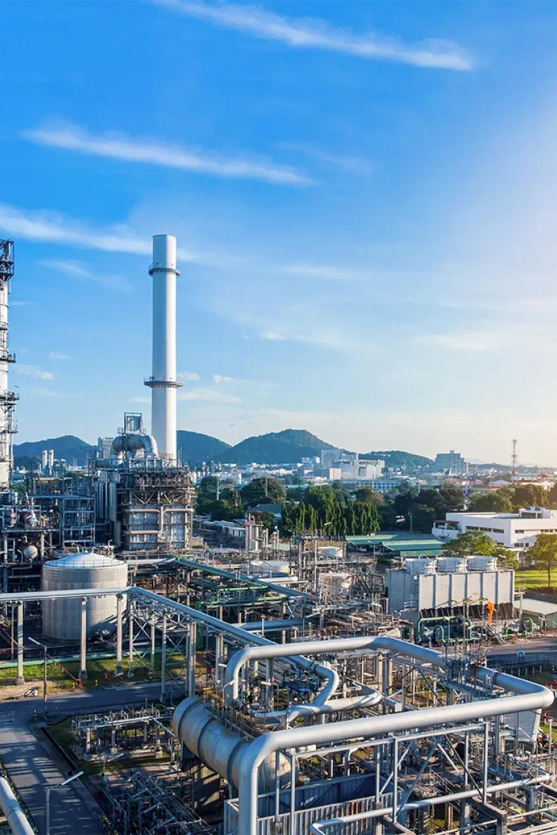 Industrial complex with pipes, storage tanks, and tall chimneys under a blue sky; mountains and a cityscape are visible in the background.