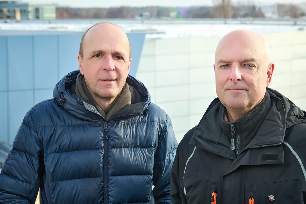 Deux hommes en veste bleue et noire devant un bâtiment industriel gris-bleu à toit plat.