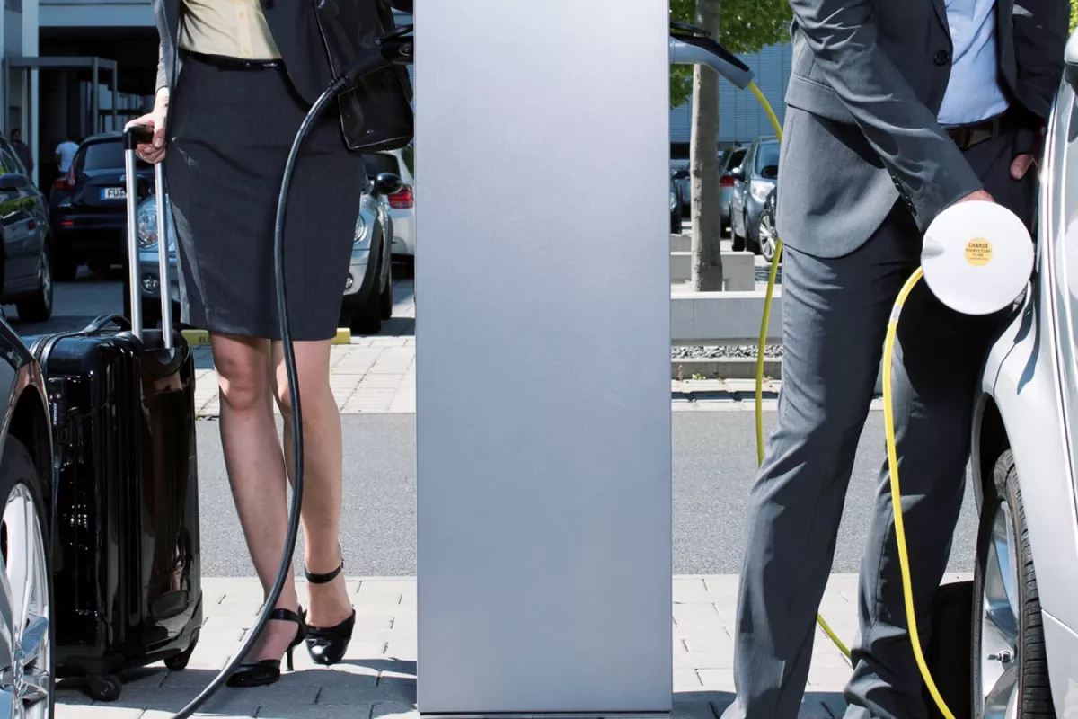 Two business professionals charging electric cars at a public ABL charging station, with luggage and office buildings in the background.