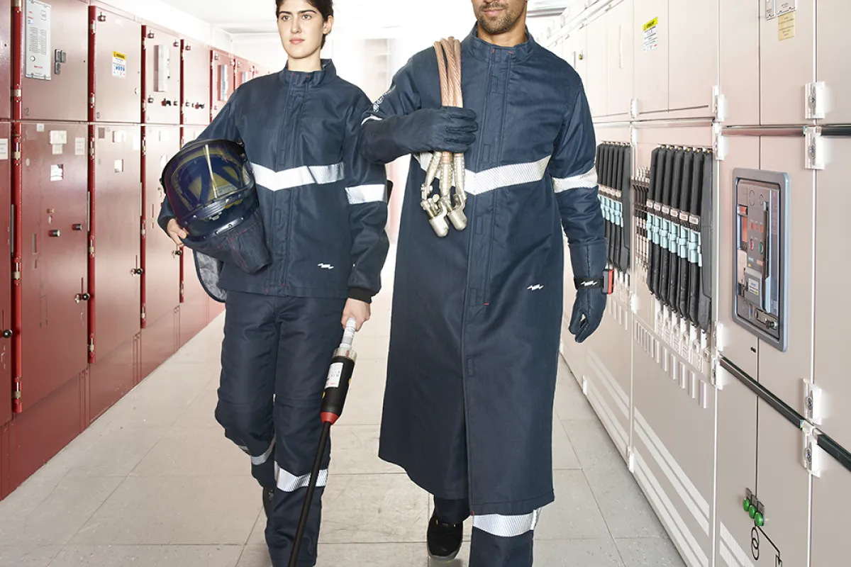 Two people in protective work uniforms walk through an industrial facility corridor, carrying tools and safety equipment.