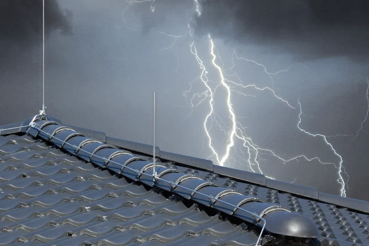 Rooftop lightning protection with metal conductors and air terminals on dark roof tiles, with lightning in a stormy sky behind.