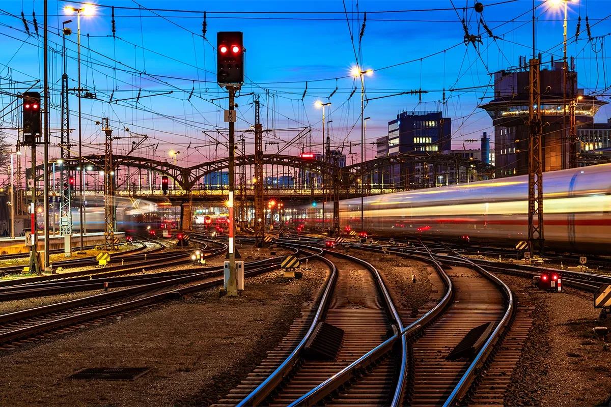 Railway junction at dusk with multiple tracks, overhead power lines, red signal lights, and blurred trains passing through an urban station area.