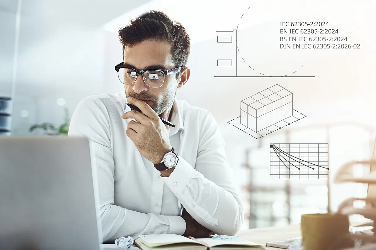 Man in glasses working at a laptop, holding a pen thoughtfully, with technical diagrams and IEC 62305-2:2024 graphics overlaid in the background.