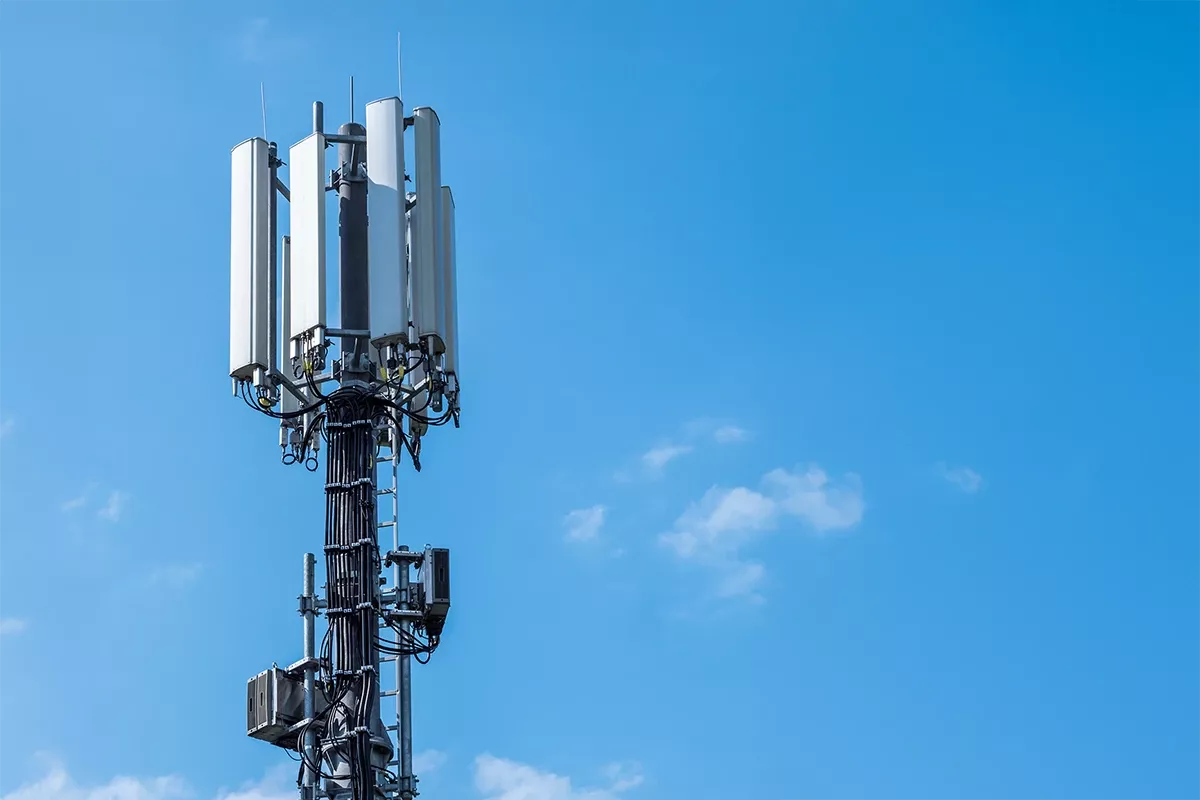 Cellular base station tower with multiple antenna panels and cables against clear blue sky.