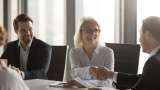 Two people are sitting at a table in an office shaking hands.